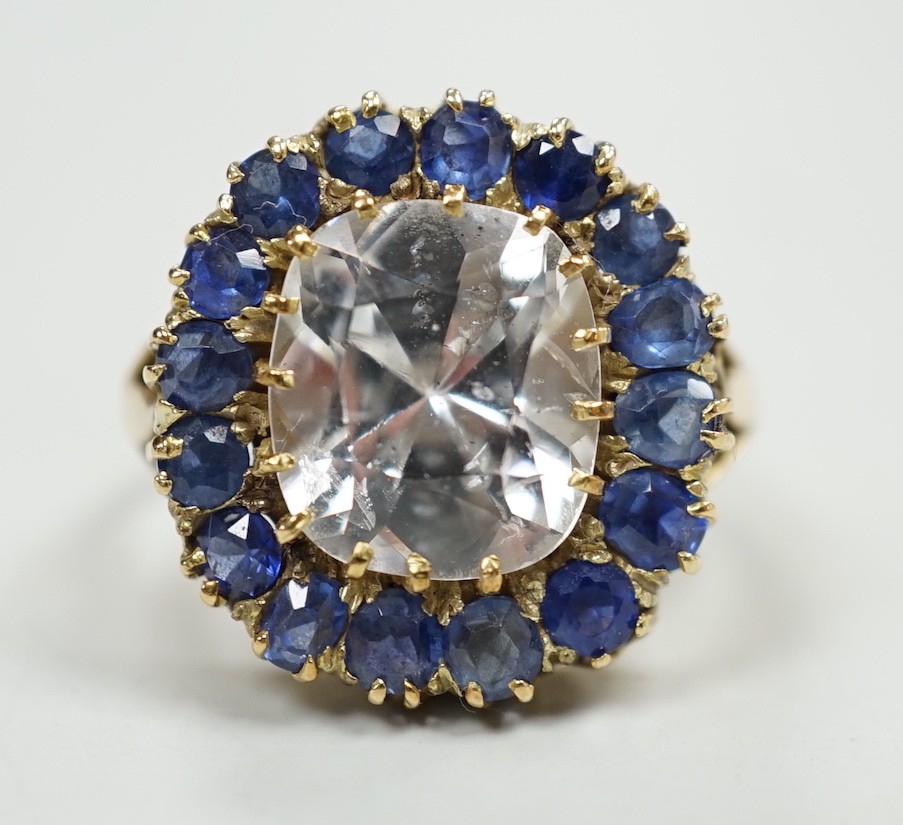 An early 20th century yellow metal and single stone cushion cut white sapphire set dress ring, bordered by round and oval cut blue sapphires, size P, gross weight 9.4 grams.